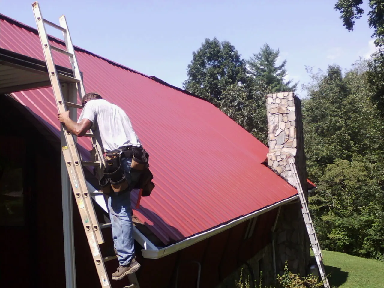 Roofer on a ladder beside a red metal roof and stone chimney — Soffit & Fascia Repair by Roofing Brothers in Fort Meade